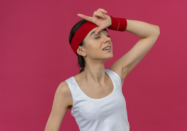 制造身穿运动服的年轻健身女士戴着头巾 手指举过头顶 闭着眼睛站在粉色的墙上做着失败者的手势姿势头带闭合
