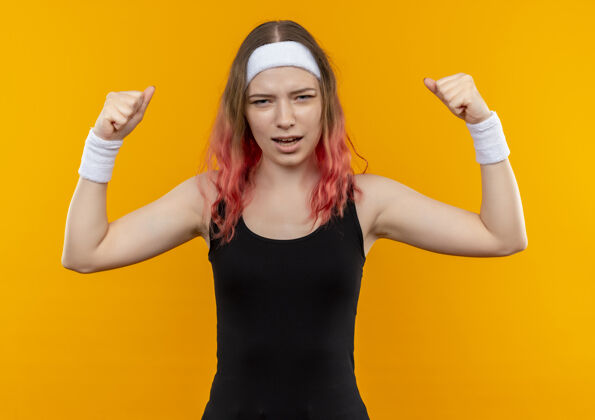 人穿着运动服的年轻健身女士站在橘色的墙上举起拳头 脸上露出恼怒的表情市民姿势健身