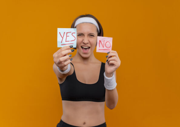 运动员身穿运动服 头箍的年轻健身女士站在橙色的墙上 兴高采烈地展示着两张写着“是”和“不是”的提醒纸纸张站立运动