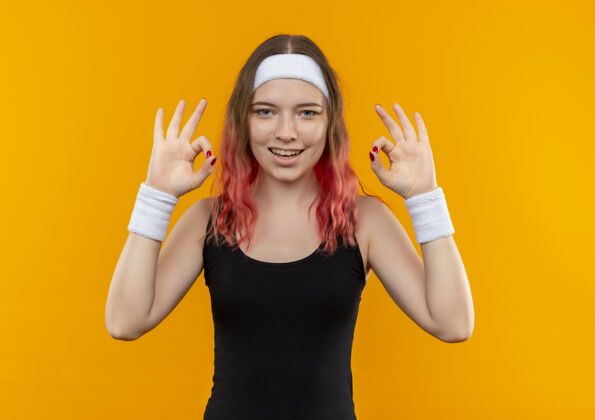 运动身着运动服的年轻健身女士 双手站在橙色的墙上 面带微笑 露出“ok”的手势欢呼手看