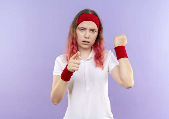 姿势年轻的运动女性用食指指着镜头握紧拳头严肃的脸站在紫色的墙上人指向运动装