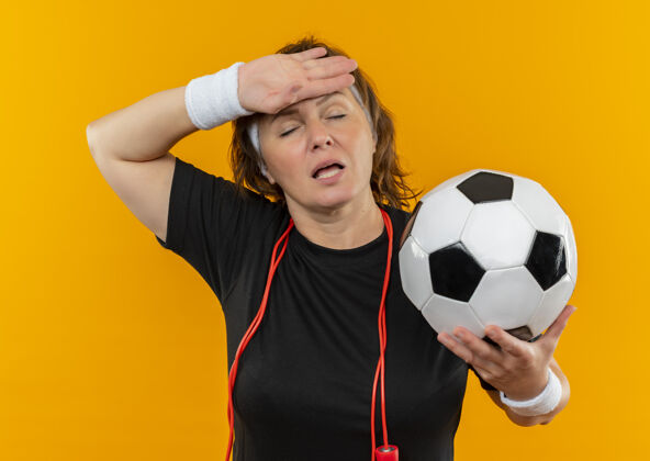 女人中年运动女性 身穿黑色t恤 头上系着足球带 站在橙色的墙上 看起来很疲倦 工作过度抱着过度工作看