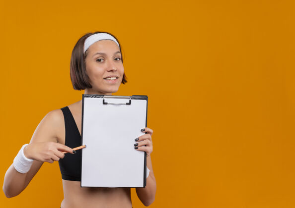 表演身穿运动服的年轻健身女士 头上戴着头带 展示着空白页的剪贴板 用笔指着剪贴板 友好地微笑着站在橙色的墙上空白健身钢笔
