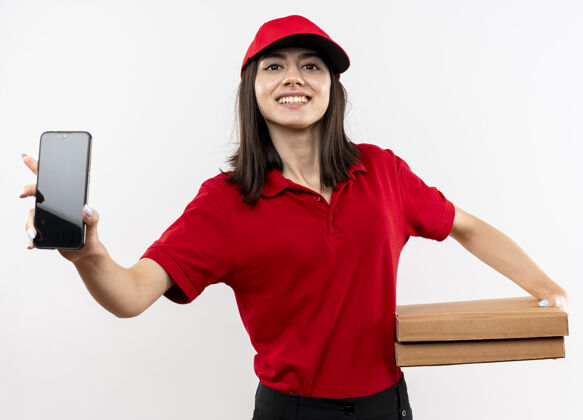 女孩身穿红色制服 头戴鸭舌帽 手持披萨盒的年轻送货女孩 展示着站在白色背景上的智能手机 开心地微笑着看着摄像机送货制服盒子