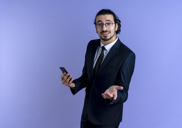 眼镜一位身穿黑色西装 戴着眼镜的商人举着智能手机 伸出手臂望着前方 站在蓝色的墙上问道公司询问优雅