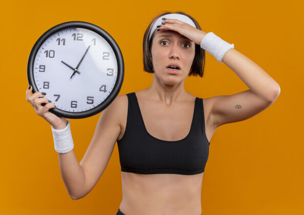 运动身穿运动服的年轻健身女 头戴头巾 手举挂钟 站在橙色的墙上 神情迷茫 非常焦虑墙非常抱着