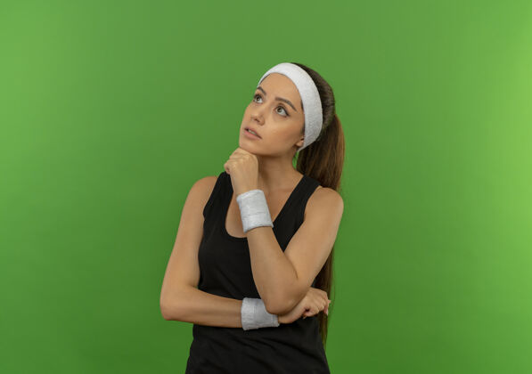 手身穿运动服的年轻健身女 戴着头巾 手放在下巴上 神情沉思地站在绿色的墙上运动下巴人