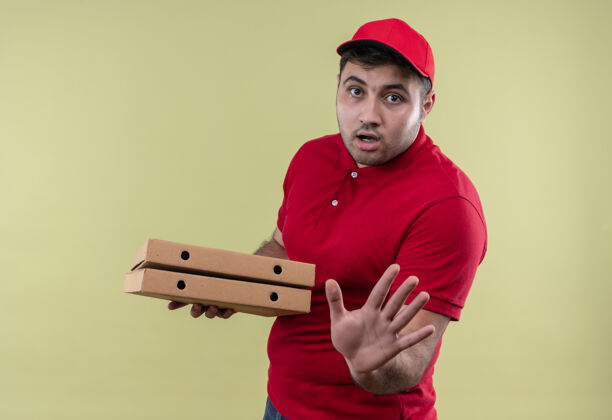 手年轻的送货员身穿红色制服 戴着帽子 手里拿着披萨盒 站在绿色的墙上 手上带着恐惧的表情做着停车标志恐惧姿势拿着