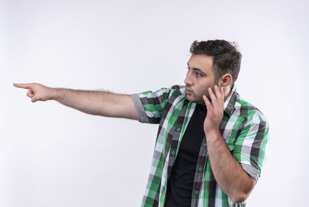 年轻穿着格子衬衫的年轻人站在白色的墙上 手指指着一边 一边用手机说话 一边显得很困惑电话指着侧线