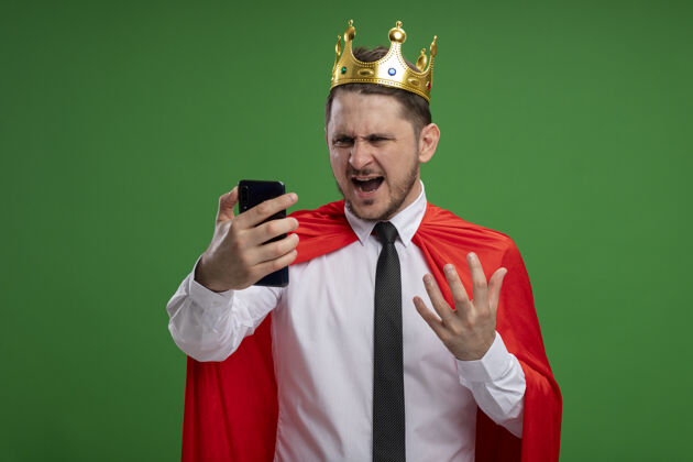 愤怒超级英雄商人穿着红色斗篷戴着皇冠用智能手机看着屏幕疯狂愤怒地站在绿色背景下屏幕斗篷野生