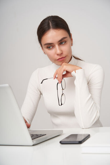 欧洲年轻漂亮的黑发女士 扎着马尾辫 坐在办公室里 用现代化的笔记本电脑 看着屏幕 面容平静 在白色的墙壁上摆姿势时 双唇合拢情感毛衣黑发