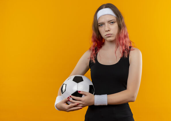 摆姿势身着运动服的年轻健身女士手拿足球 严肃的脸站在橙色的墙上球严肃运动