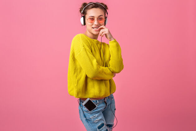 明亮迷人的快乐搞笑的调情女人戴着耳机听音乐穿着时髦的五颜六色的服装隔离在粉红色的墙上 穿着黄色的毛衣和太阳镜 玩得很开心时尚学生音乐