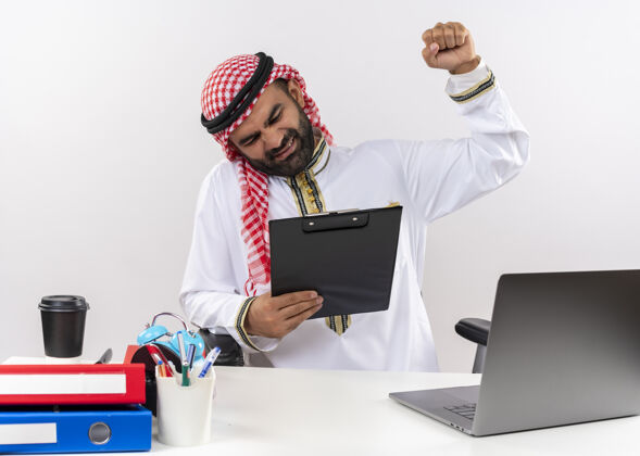穿身着传统服装的阿拉伯商人坐在桌旁 手持笔记本电脑 握紧写字板 表情烦躁地握紧拳头在办公室工作剪贴板拳头工作