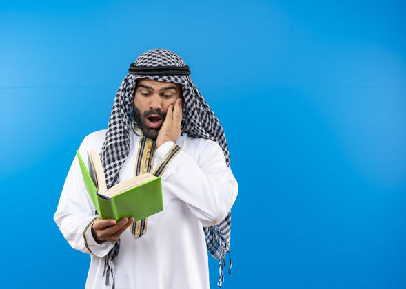 震惊一个穿着传统服装的阿拉伯人站在蓝色的墙上看着一本书 震惊不已穿传统男人