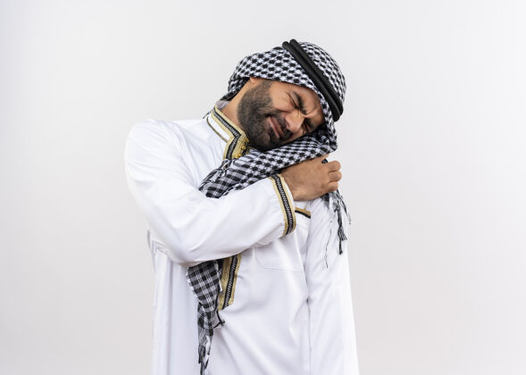 传统一个穿着传统服装的阿拉伯男人站在白墙上 看着不舒服 摸了摸肩膀 感到疼痛碰男人疼痛