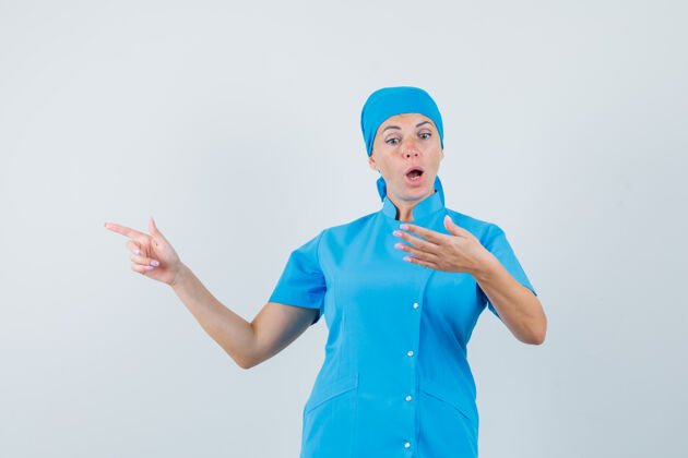 年轻人女医生假装拿着手机 穿着蓝色制服指着一边 惊讶地看着前视图疾病指点女人