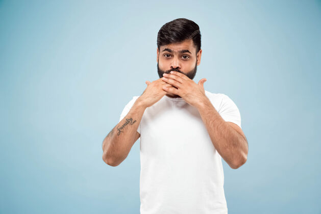 表情蓝色背景上穿着白衬衫的印度教年轻人的半身特写肖像人类情感 面部表情 销售 广告概念负面空间用手捂住脸工作年轻男性