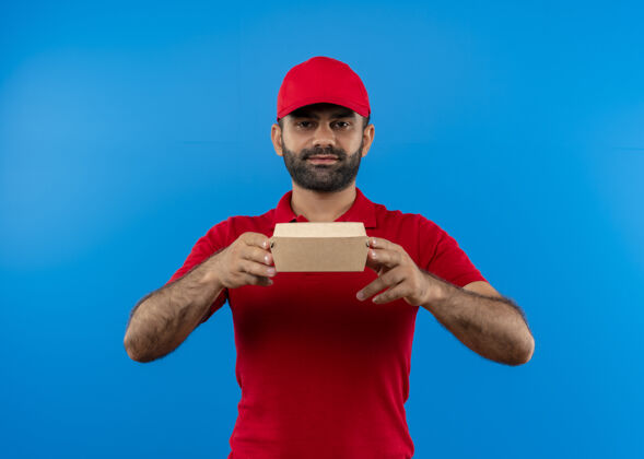 立场留着胡须的送货员身着红色制服 戴着帽子 站在蓝色的墙上 露出自信的微笑 展示着盒子包装信心微笑制服