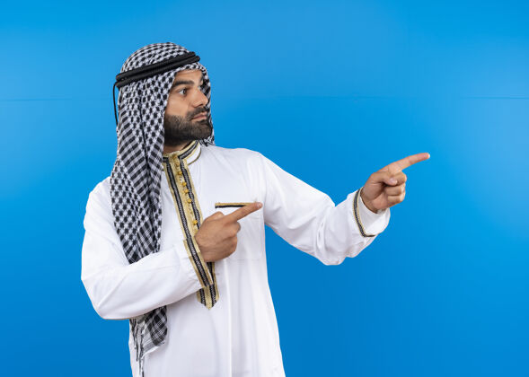 看身着传统服装的阿拉伯男子一边看一边用手指指着一边 严肃的脸站在蓝色的墙上手指站严重