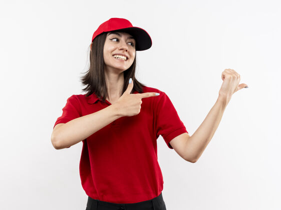 帽子身穿红色制服 头戴鸭舌帽的年轻送货女 手指指向侧面 站在白色背景下 脸上露出幸福的笑容站微笑制服