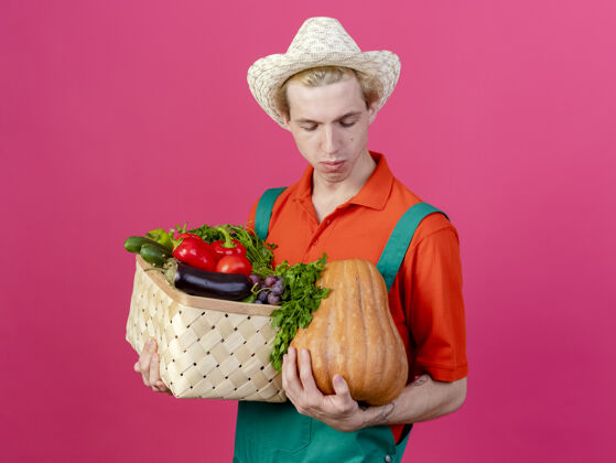 抱着年轻的园丁穿着连体衣戴着帽子拿着装满蔬菜的箱子蔬菜穿下来