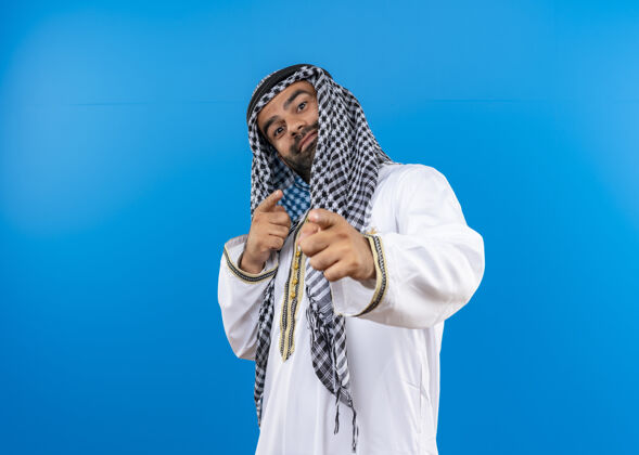 手指一位身着传统服装的阿拉伯男子站在蓝色的墙上 用手指着脸 脸上带着自信的微笑男人传统穿着