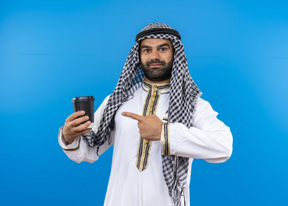 脸身着传统服装的阿拉伯男子站在蓝色的墙上 用手指指着智能手机 脸上带着微笑阿拉伯语手指指着