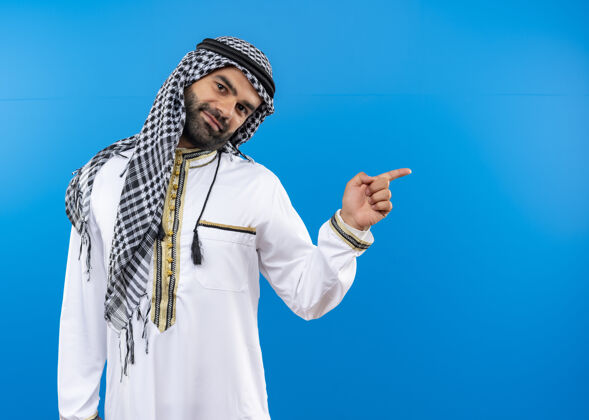站身着传统服装的阿拉伯男子 脸上带着微笑 手指着站在蓝色墙上的一边阿拉伯语侧线手指