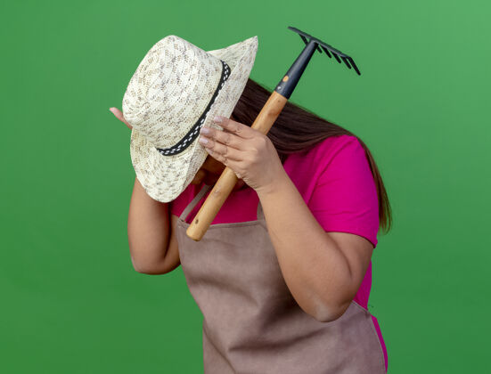 无聊穿着围裙戴着帽子拿着迷你耙子的中年园丁女人女人花园站着