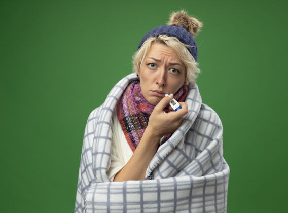 包裹生病的不健康的妇女 短头发 戴着暖和的围巾 戴着帽子 感觉不舒服 裹着毯子 嘴里叼着温度计 站在绿色的背景上嘴帽子体温计