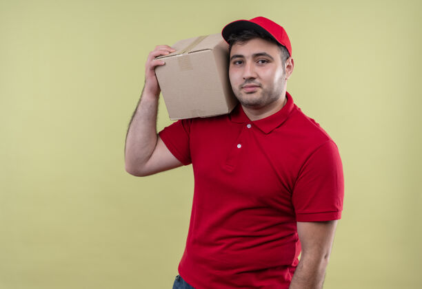 人年轻的送货员穿着红色制服 戴着帽子 拿着盒子 微笑着自信地站在绿色的墙上帽子公民微笑