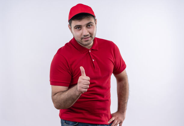 拇指年轻的送货员身穿红色制服 戴着帽子 站在白色的墙上 愉快地笑着竖起大拇指年轻人微笑男人
