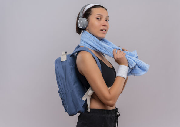 健身年轻的健身女士穿着运动服 背着背包 头戴着耳机 脖子上戴着毛巾 站在白色的墙上 她看起来自信地微笑着自信脖子健康