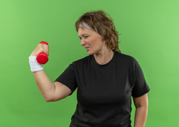 健康中年运动女性 身穿黑色t恤 戴着头带 举着哑铃 自信地站在绿色的墙上中间女人站