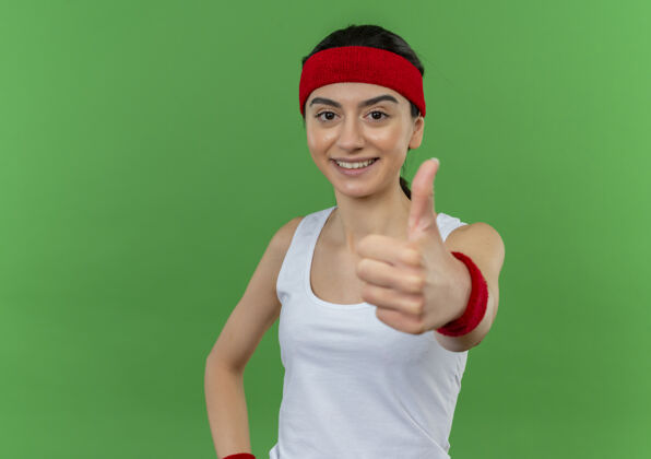 市民身穿运动服的年轻健身女士 头戴花环 快乐而积极地微笑着 站在绿色的墙上 愉快地竖起大拇指看欢呼表演