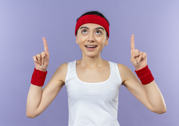 健身身穿运动服的年轻健身女士 戴着头带微笑 自信地用食指指着紫色的墙壁女人运动装摆姿势