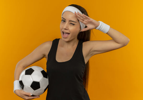 运动员身穿运动服 头戴足球带的年轻健身女士站在橙色的墙上 微笑着眨眼致意女人人市民