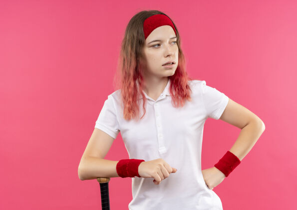 运动戴着头巾 拿着球棒 严肃的脸站在粉色墙壁上的年轻运动女郎女人女人摆姿势