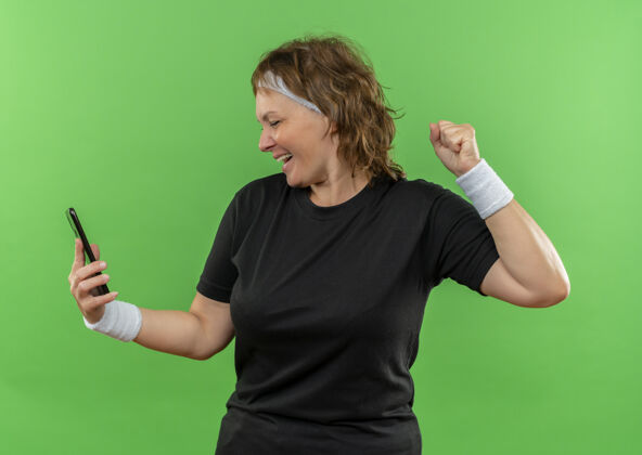 人中年运动女性 身穿黑色t恤 戴着头巾 看着手机屏幕 握紧拳头 站在绿色的墙上 快乐而兴奋握紧兴奋头带