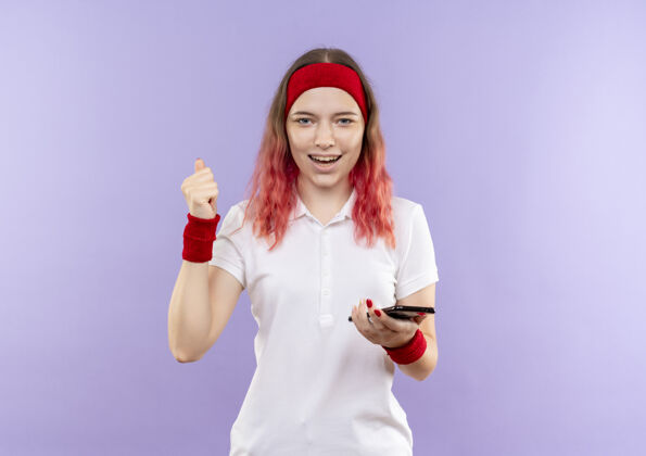 女人年轻的运动型女子手持智能手机握紧拳头 站在紫色的墙壁上愉快地微笑着人市民移动