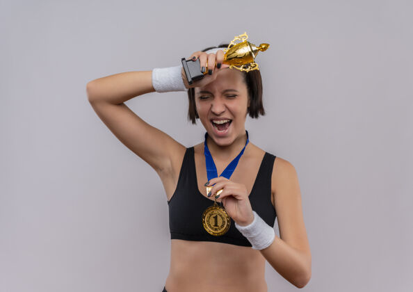 人一位身穿运动服 脖子上挂着金牌 手持奖杯的年轻健身女士站在白墙上为自己的成功感到高兴和兴奋抱着成功女