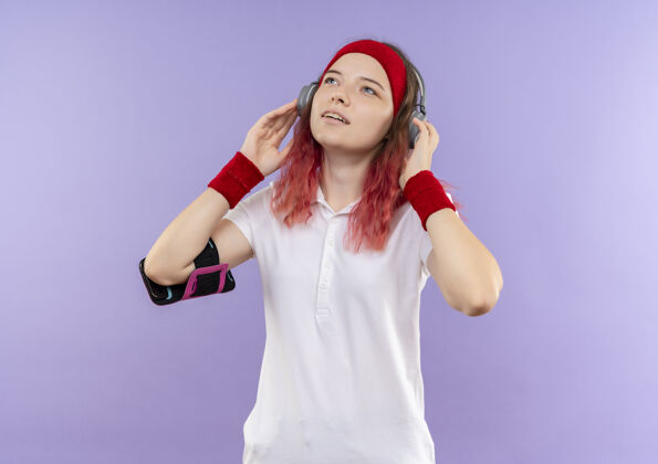 音乐戴着头戴耳机的年轻运动女性抬头欣赏着她最喜欢的音乐 手持智能手机袖标站在紫色的墙上训练年轻长相人物