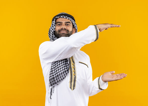 传统身着传统服装的阿拉伯商人站在橙色的墙上 脸上挂着大大的标志 微笑 测量符号测量阿拉伯语站立