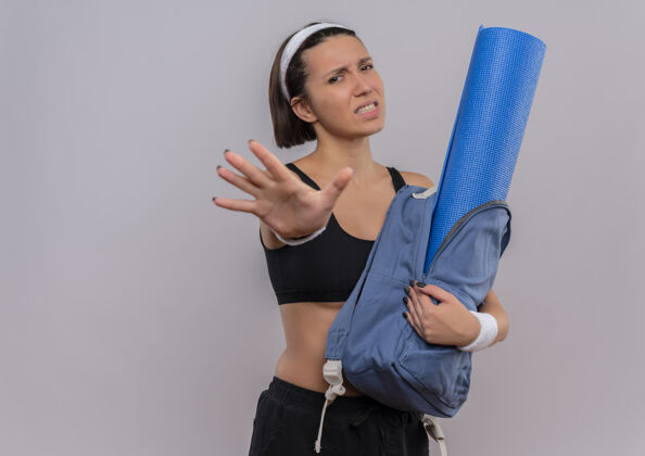 人穿着运动服的年轻健身女士拿着背包 手拿瑜伽垫 手张开 站在白色的墙上 表情厌恶地做着停车标志运动员垫子女人