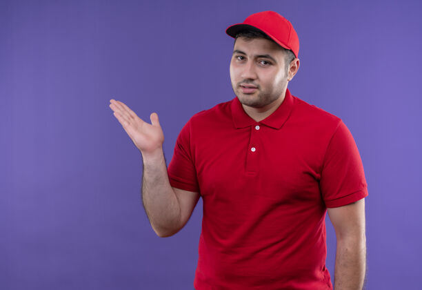交货年轻的送货员身穿红色制服 戴着帽子 手举手臂站在紫色的墙上 展示复印空间人人人