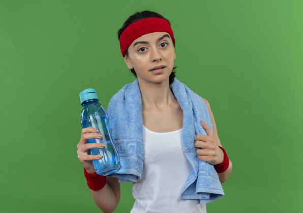 健身一位身穿运动服的年轻健身女士 头上戴着头巾 脖子上挂着毛巾 手里拿着一瓶水 站在绿色的墙上 看上去很疲惫水市民头带