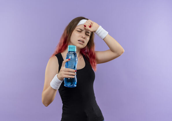 姿势穿着运动服的年轻健身女士站在紫色的墙上 手里拿着一瓶水 看上去很不舒服 头痛得厉害强壮人运动