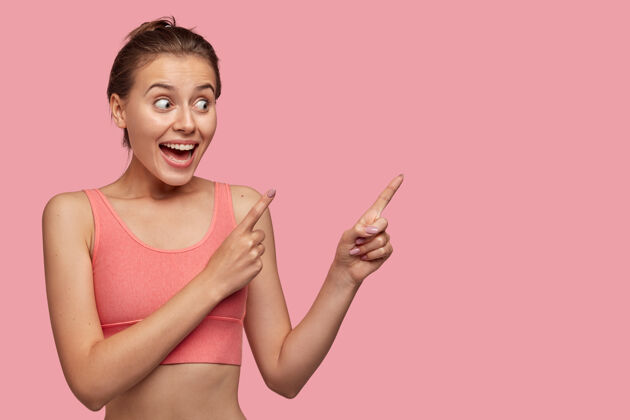 展示身材完美的漂亮年轻女子的水平镜头 表示食指放在一边 很高兴展示一些物品 隔离在粉红色的墙上女运动员宣传装备女运动员牙齿旁边