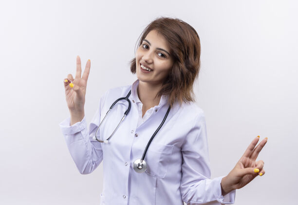 表演身穿白大褂 手持听诊器的年轻女医生微笑着 脸上挂着胜利的标志 站在白墙上站立脸外套